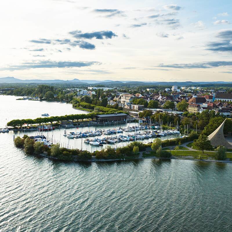 Luftaufnahme Radolfzeller Hafen mit vielen Segelbooten, umgeben von einer grünen Landschaft und die Stadt und das Münster im Hintergrund.