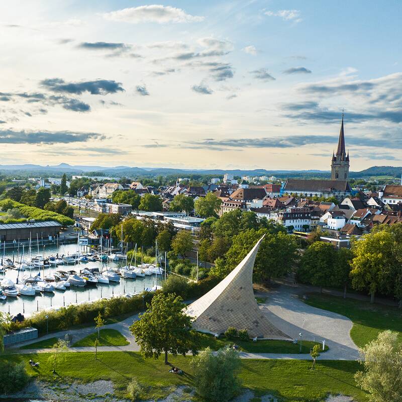 Die Luftaufnahme zeigt die Mole mit vielen Booten und dem Konzertsegel im Vordergrund und die Stadt Radolfzell mit Münster im Hintergrund, umgeben von grünem Landschaftsgebiet.