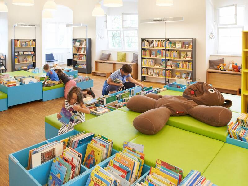 Ein bunter Bibliotheksbereich für Kinder mit Büchern und einem großen Stofftier, in dem mehrere Kinder lesen und spielen.