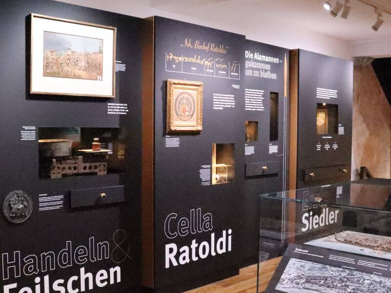 Ein Ausstellungsraum mit schwarzen Wänden, auf denen verschiedene historische Exponate, Texte und Bilder zur Geschichte Radolfzells präsentiert werden.