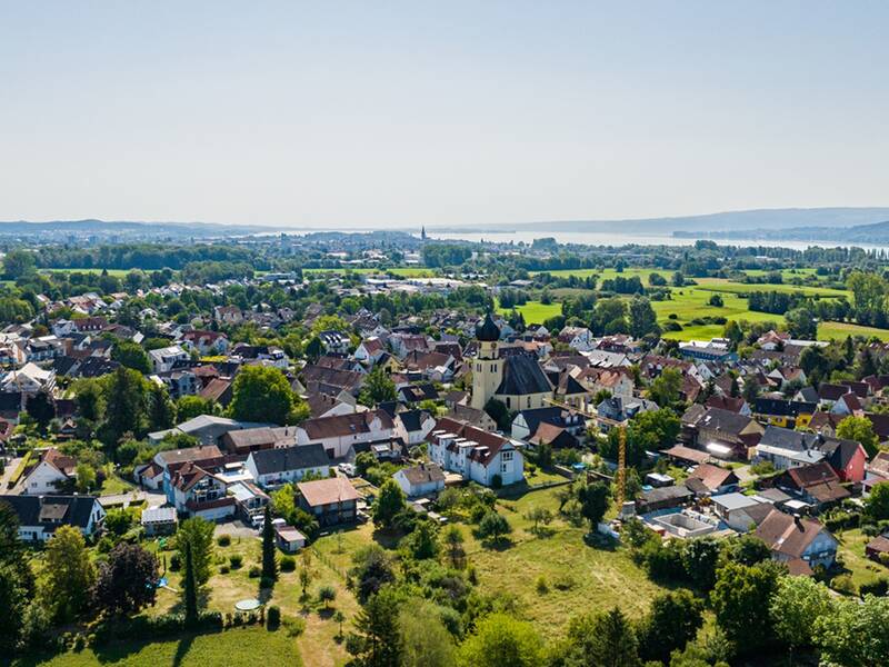 Eine malerische Luftaufnahme zeigt Böhringen mit der Kirche St. Nikolaus, umgeben von grünen Feldern und dem Bodensee im Hintergrund.