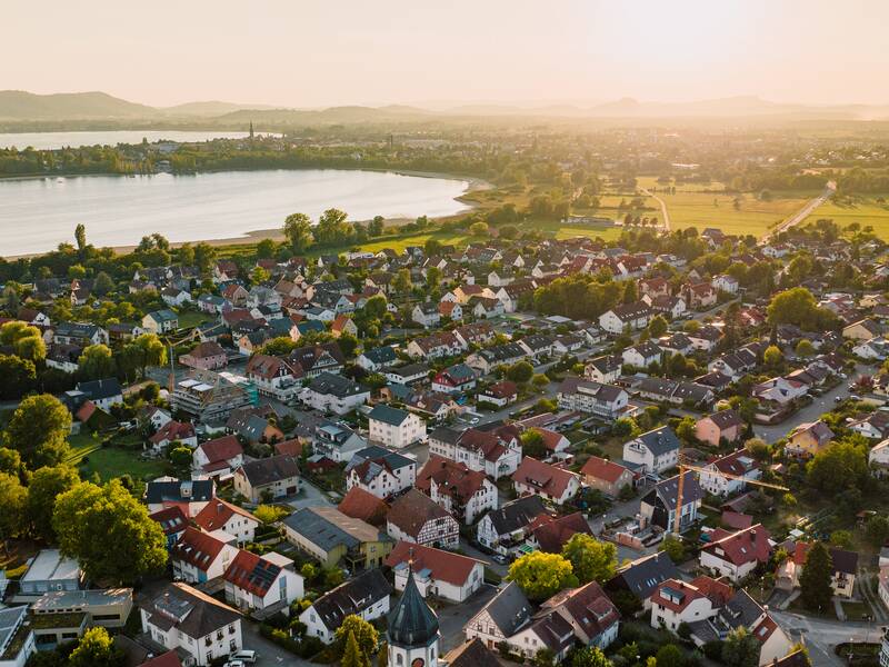 Luftaufnahme von Markelfingen mit Häusern, Bäumen und dem Bodensee im Hintergrund bei Sonnenuntergang.