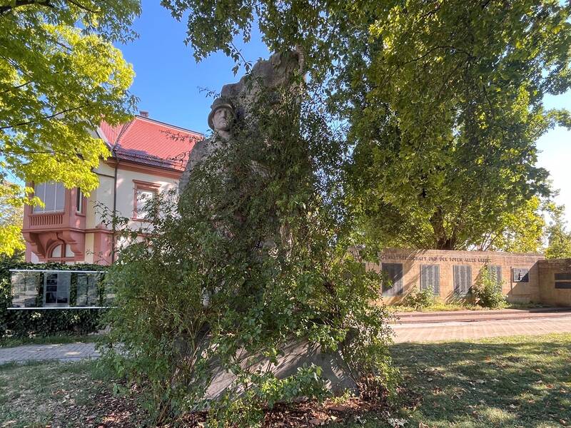 Das NS-Kriegerdenkmal am Luisenplatz teilweise überwuchert von einer Rose, steht in einem parkähnlichen Bereich vor einem rosafarbenen Gebäude mit rotem Dach, umgeben von Bäumen.