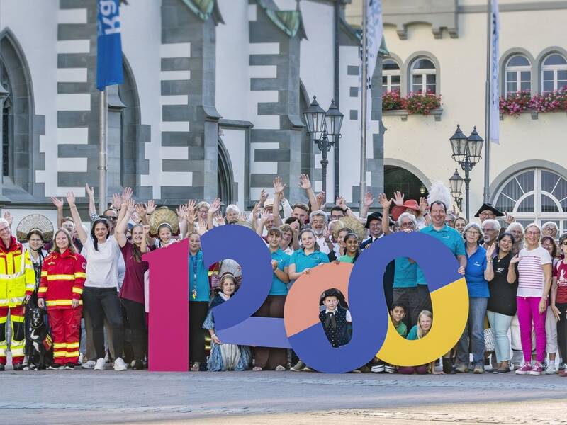 Eine große Gruppe von Menschen steht vor dem Radolfzeller Rathaus  hinter einer bunten Skulptur der Zahl 1200, einige tragen Uniformen und fröhlich die Hände in die Luft.