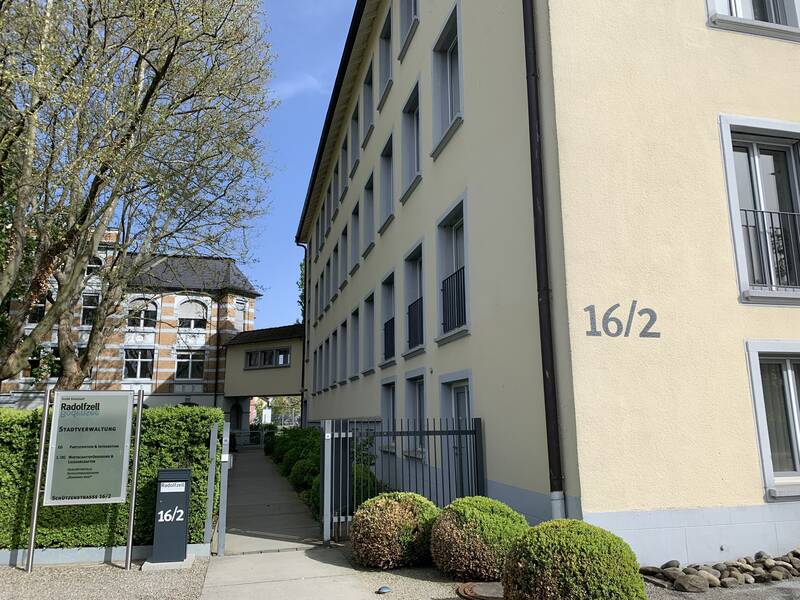 Das hellgelbe Dienstgebäude mit der Nummer 16/2 neben einem großen, blühenden Baum und einem Schild, das auf die Stadtverwaltung hinweist.