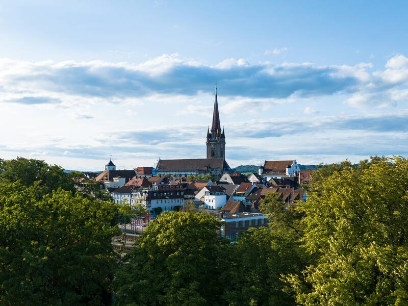 Radolfzell mit dem Münsterturm in der Mitte, umgeben von Häusern und Bäumen unter einem blauen Himmel mit vereinzelten Wolken.