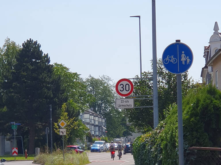 Eine Straße mit Verkehrsschildern, darunter ein Tempolimit von 30 km/h und ein Schild getrennter Geh- und Radweg, neben einem Baum und Wohnhäusern.