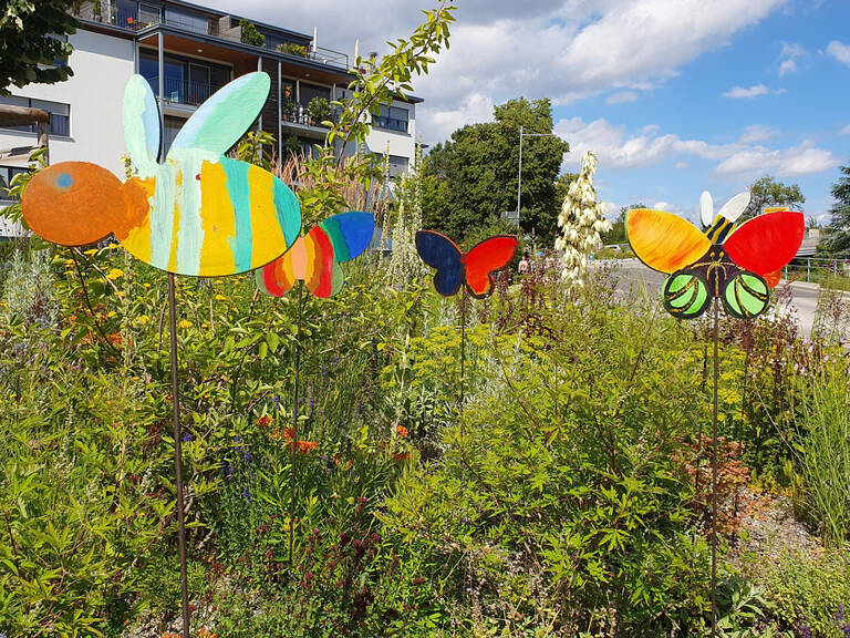 Bunte, aus Holz gefertigte Schmetterlings- und Bienenfiguren stecken auf Stäben in einer Wiese vor einem Wohngebäude.