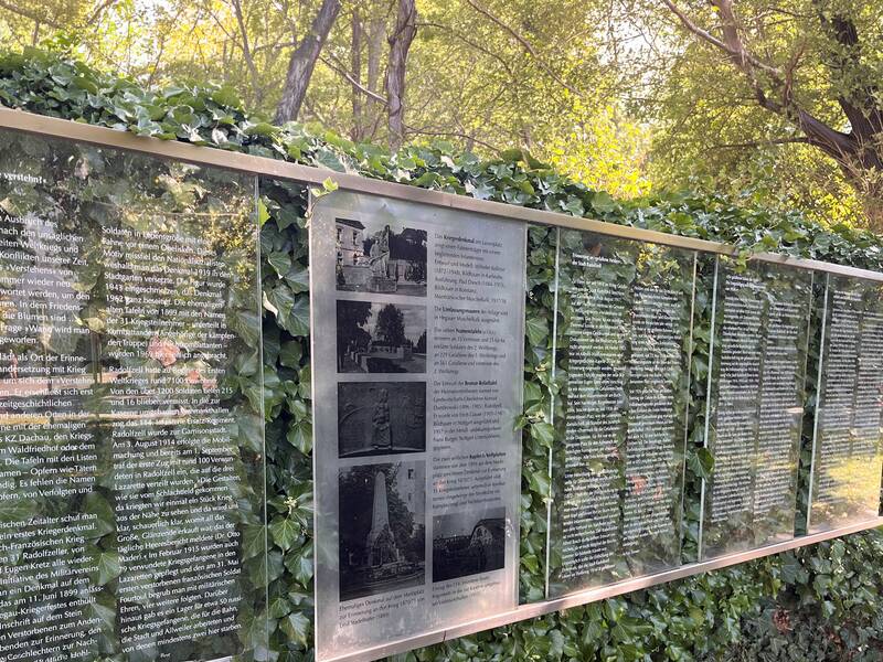 Die Tafeln am Kriegerdenkmal mit Texten und Bildern stehen in einer Umgebung, die von grünem Efeu bewachsen ist.