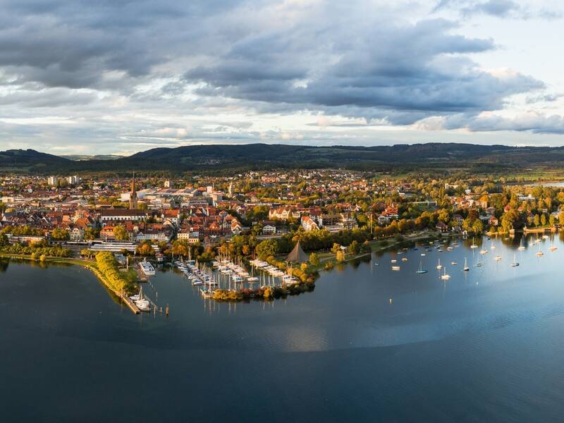 Luftaufnahme von Radolfzell  mit Segelbooten im Hafen und Hügeln im Hintergrund unter einem bewölkten Himmel.