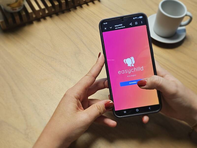 Eine Frau hält ein Smartphone mit einer geöffneten App namens "easychild". Im Hintergrund steht eine weiße Tasse auf einem Holztisch.