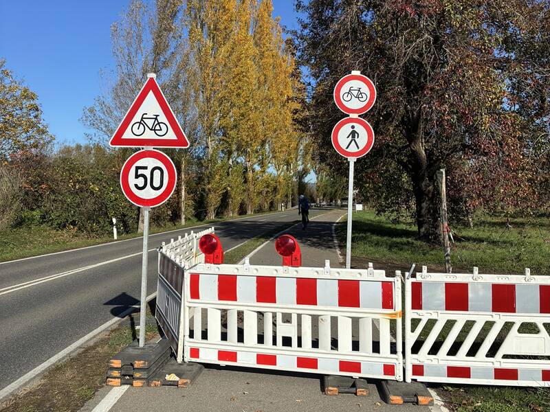 Straße und Radweg sind durch Absperrungen und Verkehrsschilder für Radfahrer und Fußgänger gesperrt, während ein einzelner Radfahrer auf der sonnigen Strecke fährt.