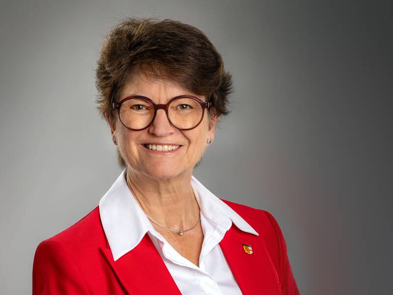 Eine lächelnde Bürgermeisterin Monika Laule trägt eine Brille und einen roten Blazer über einer weißen Bluse vor einem grauen Hintergrund.