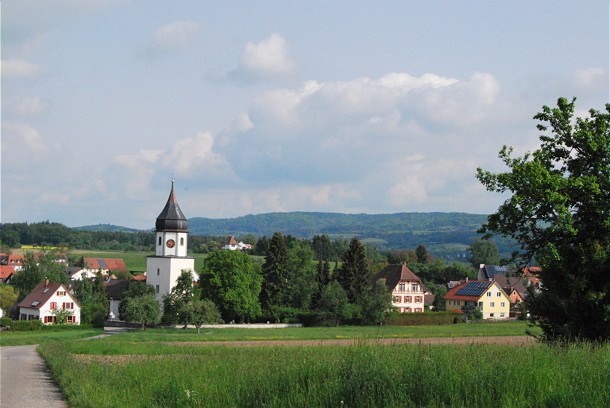  Ansicht von Markelfingen, im Vordergrund eine Wiese, im Hintergrund sieht man die Kirche und Häuser 