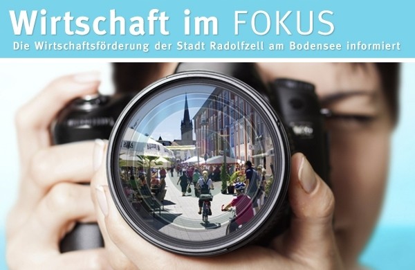  Person mit Kamera, in der Linse sieht man Radolfzell mit Fußgängern und Fahrradfahrern, im Hintergrund ist das Münster zu sehen 