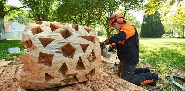  Großer Baumstamm ohne Rinde, mit vielen rechteckigen und dreieckigen Auskerbungen, Person mit Schutzkleidung und Motorsäge bearbeitet ihn, Bild: Kuhnle und Knödler 
