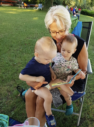  Frau im Gartenstuhl hat zwei Kinder auf dem Schoß, alle drei schauen auf ein Buch in der linken Hand der Frau 